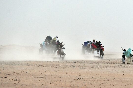 Arlit, près de 1200 km de Niamey, capitale du Niger. Cette ville célèbre pour son uranium est aussi une zone de transit de certains migrants en partance pour l’Europe ou les pays maghrébins comme l’Algérie ou la Libye. Une femme et des enfants saluent les migrants assis dans des pick-up, tenant des bâtons de bois attachés au véhicule pour éviter de tomber, alors qu'ils quittent la banlieue d'Agadez. Agadez le 1er juin 2015. PHOTO AFP / ISSOUF SANOGO ISSOUF SANOGO / AFP